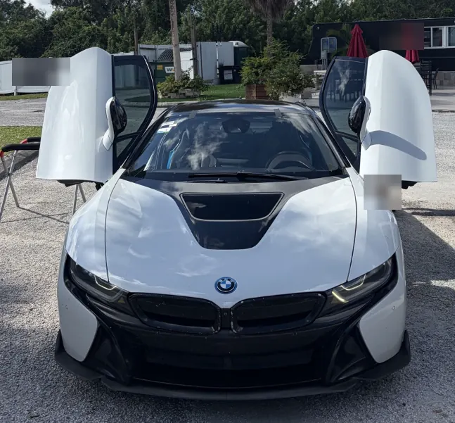 White BMW i8 with its butterfly doors open, parked in a gravel lot for windshield replacement