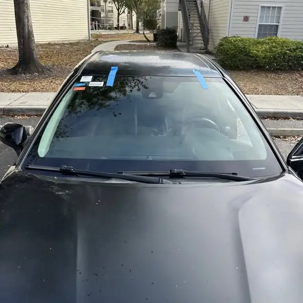 Newly installed windshield, still held by blue retention tape, on a dark sedan parked outside a customer's home