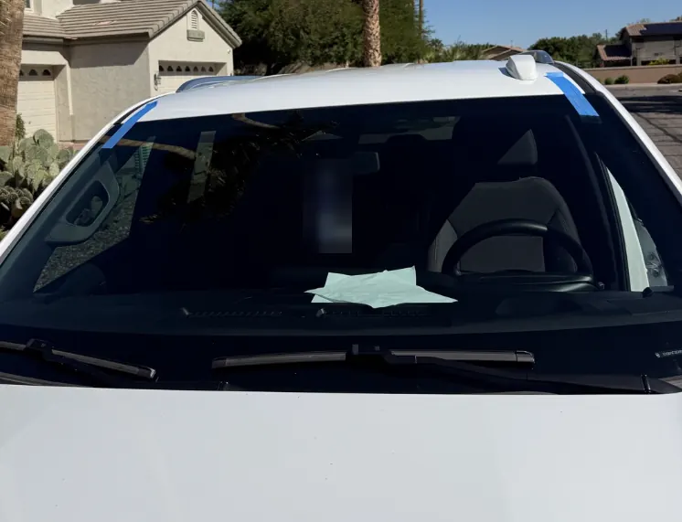 White car with a freshly installed windshield, blue retention tape on the corners, outside a stucco home with cactus in Arizona